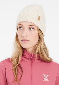 Crème gebreide beanie met een gestructureerd patroon en een klein bruinen leren logo label. Gekleed in een roze ritsjack met witte accenten.