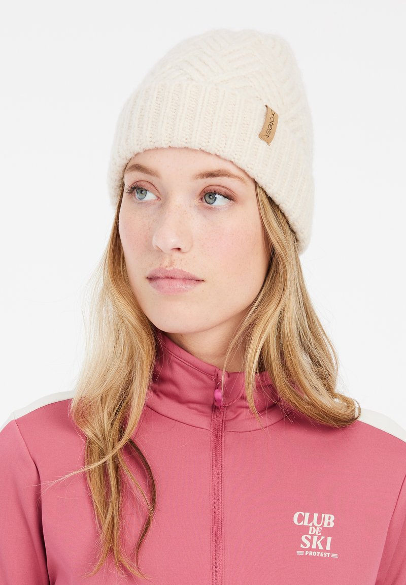 Crème gebreide beanie met een gestructureerd patroon en een klein bruinen leren logo label. Gekleed in een roze ritsjack met witte accenten.