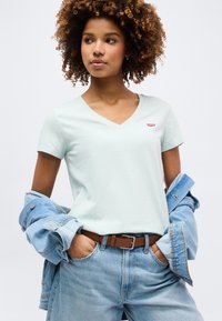 Νεαρή γυναίκα με κατσαρά μαλλιά που φοράει ανοιχτό μπλε T-shirt με V, τζιν μπουφάν περασμένο στους αγκώνες, μπλε τζιν και καφέ ζώνη.