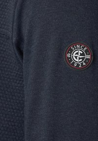 Donker marineblauwe sweatshirtstof met een gestructureerd patroon, voorzien van een ronde, rood en zwarte badge met de tekst "SINCE 1924" in de linkerbovenhoek.