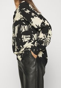 Chemise  à manches longues noire avec un motif floral crème, confectionnée dans un tissu léger. Associée à un pantalon en cuir noir, les mains dans les poches.