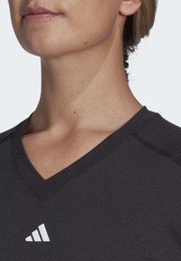 Camiseta deportiva negra de cuello en V, hecha de una tela suave, con un logo blanco en el lado izquierdo del pecho y costuras reforzadas en los bordes.