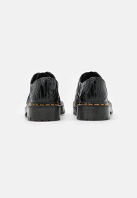 Černé boty s lesklým povrchem, které mají vlnitou texturu, zaoblený podpatek a žluté prošívání podél podešve. Tlustá, vroubkovaná podešev.