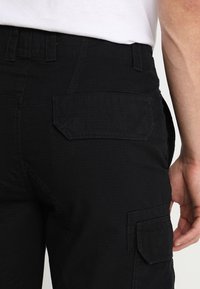 Gros plan sur une personne portant un pantalon cargo noir avec des poches à rabat à l'arrière et sur les côtés, associé à une chemise blanche.