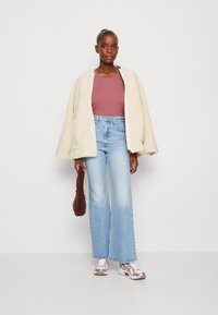 Chaqueta de forro polar crema de gran tamaño, top ajustado color malva, jeans de campana azul claro, zapatillas gris y blancas, sosteniendo un bolso texturizado marrón.