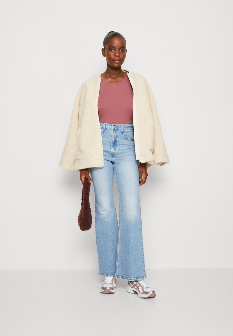 Chaqueta de forro polar crema de gran tamaño, top ajustado color malva, jeans de campana azul claro, zapatillas gris y blancas, sosteniendo un bolso texturizado marrón.