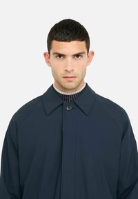 Cappotto blu scuro con ampio colletto, chiusura con bottoni e maniche raglan; texture liscia e design minimalista. Il materiale esterno è leggero.