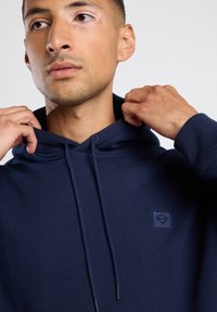 Sudadera azul marino de tela suave, con un bolsillo canguro en la parte delantera, cordones y un pequeño parche con el logo en el pecho.