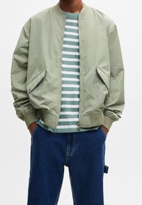 Giacca bomber verde chiaro con chiusura a zip, tasche laterali e polsini a coste, indossata sopra una camicia a righe blu chiaro e bianco e jeans di denim.