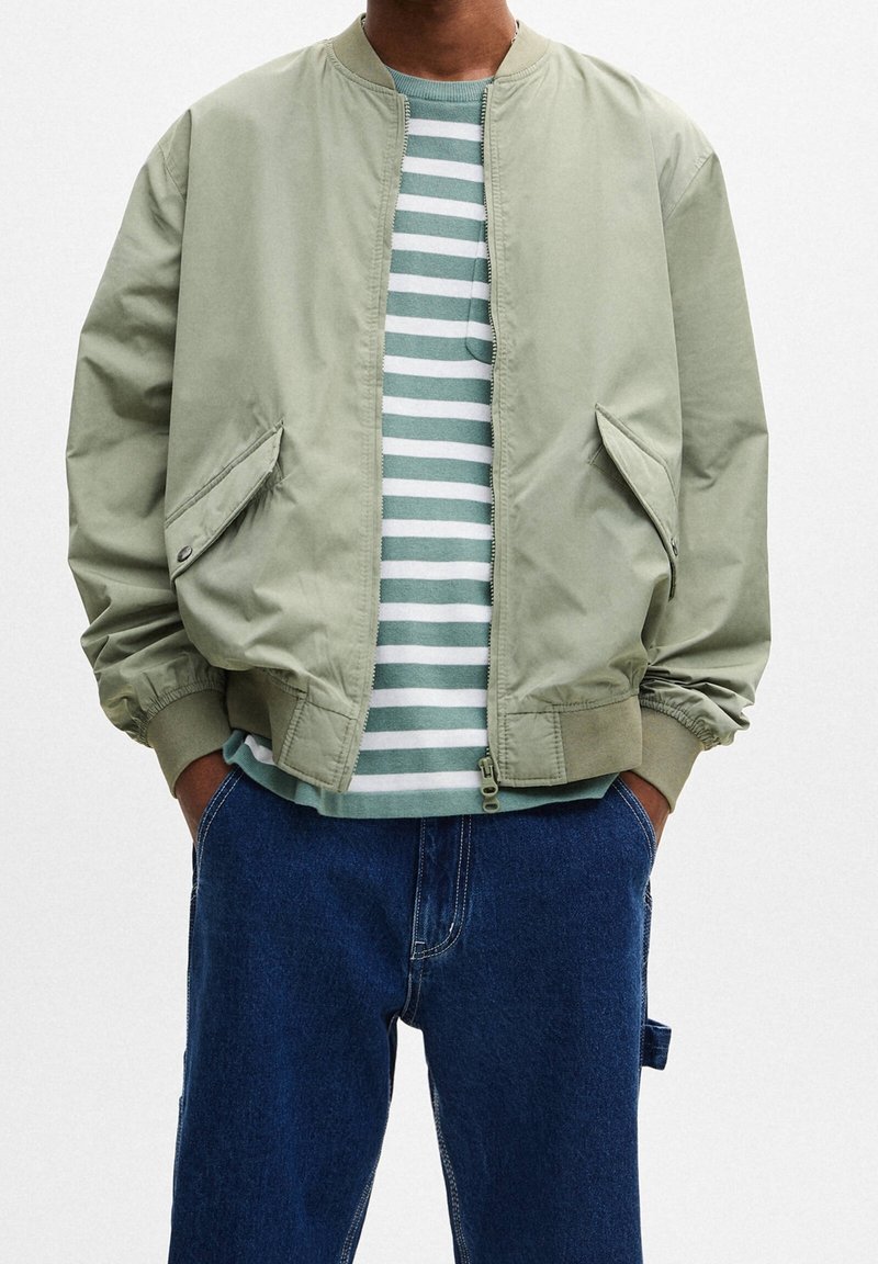 Giacca bomber verde chiaro con chiusura a zip, tasche laterali e polsini a coste, indossata sopra una camicia a righe blu chiaro e bianco e jeans di denim.