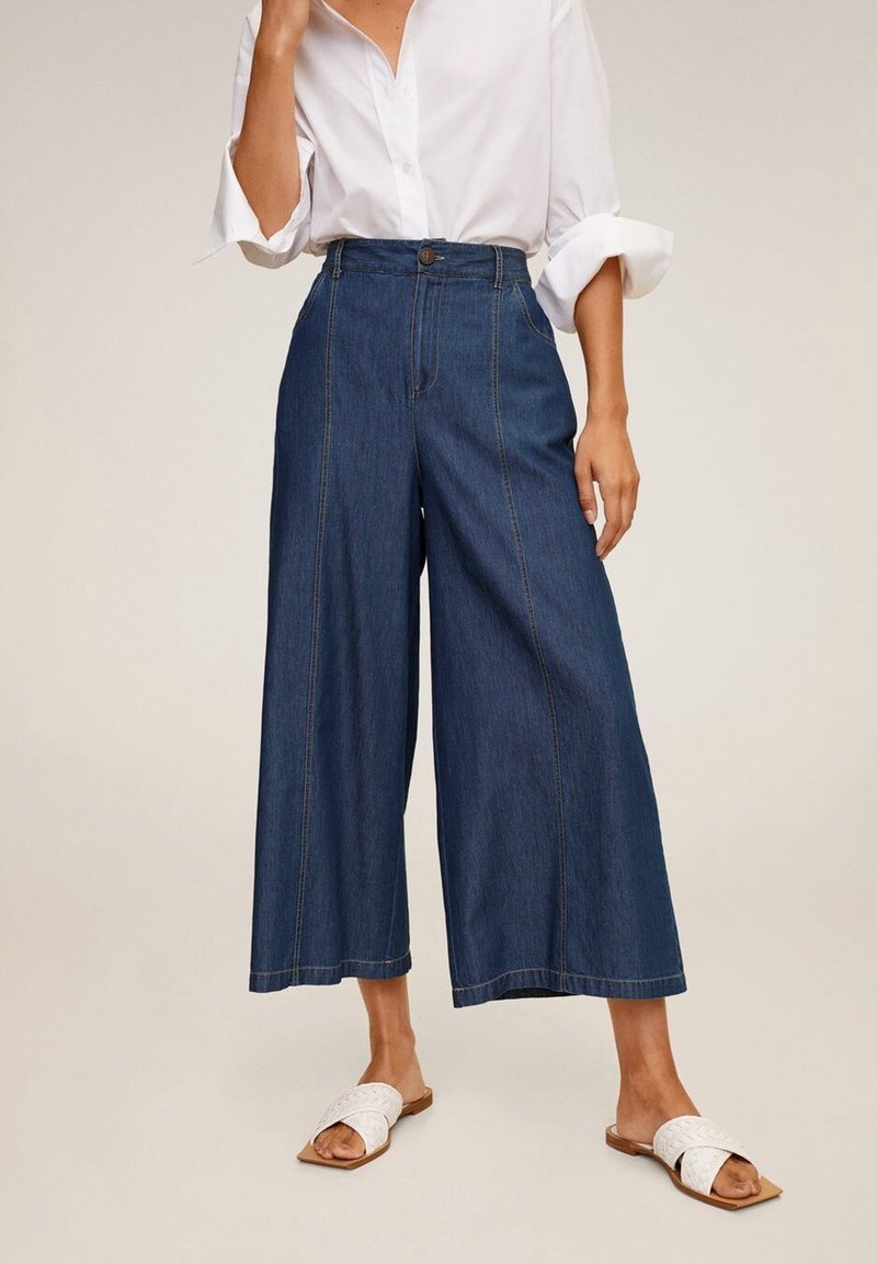 Culottes en denim à jambe large de couleur bleu foncé avec une taille haute, présentant des détails de couture visibles et une fermeture à bouton. Associés à des sandales plates blanches.