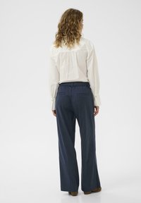 Göndör hajú nő áll háttal, fehér blúzt visel, amelyet kék csíkos, bő szárú nadrágba tűrt és barna cipőt visel.