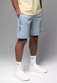 Shorts de sport bleu clair avec poches latérales, arborant une impression de texte noir sur la jambe gauche. Portés avec des baskets blanches et des chaussettes blanches côtelées.