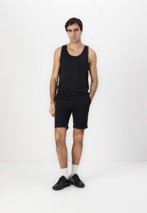 Schwarzes Tanktop und schwarzes Shorts-Outfit. Das Material scheint Baumwolle zu sein. Mittellange Shorts mit Seitentaschen. Schwarze Sneakers und weiße Socken.