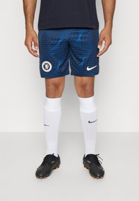 Pantalones cortos azules con la marca del Chelsea FC, con un patrón de cuadrícula, parche de logo de goma, combinados con calcetas blancas hasta la rodilla y botas negras.