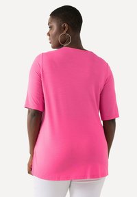 Roze top met korte mouwen en een ronde halslijn, gemaakt van zachte stof met een ontspannen pasvorm, de zoom valt onder de heupen. Zilveren oorbellen in de vorm van ringen.