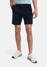 Mörkblå shorts i mjukt material med rak skärning, sidofickor och rullade fållar. Bärs med grå och vita löparskor.