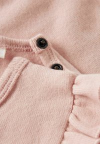 Šviesiai rožinė apranga, turinti tekstūrinį mezgimą, su juodais spaudžiamaisiais mygtukais ir raukšlėmis kraštų. Artimas audinio detalės planas.