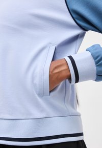 Χέρι με σκούρο τόνο δέρματος που ακουμπά μέσα στην τσέπη ενός ανοιχτόμπλε μπουφάν, το τριμ της μανσέτας και του τελικού μέρους του μπουφάν είναι με μαύρες και άσπρες ρίγες.