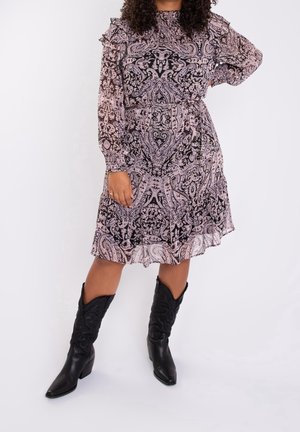 Vrouw draagt een lange jurk met lange mouwen in roze en zwart paisley dessin met ruches, gecombineerd met zwarte cowboylaarzen tot halverwege de kuit.