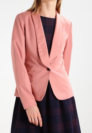 Blazer rose avec une coupe ajustée, revers crantés, fermeture à un bouton et manches longues, associé à une robe à carreaux foncés.