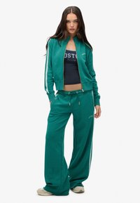 Felpa verde e pantaloni da jogging abbinati in tessuto morbido. La felpa ha una chiusura con zip e strisce bianche a contrasto; i pantaloni presentano una vita con coulisse.