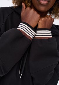 Czarna bluza z kapturem, z regulowanym sznurkiem, z paskowanymi, prążkowanymi mankietami w kolorze białym, granatowym i brązowym. Materiał wydaje się miękki o gładkiej teksturze.