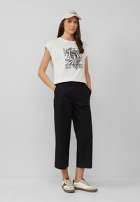 Witte graphic t-shirt met zwarte print, zwarte cropped broek en beige sneakers met bruine accenten. Een lichte pet maakt de outfit compleet.