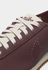 Κοντινό πλάνο μπορντό αθλητικού παπουτσιού Nike με λευκά κορδόνια, διατρητό σχέδιο swoosh και τη λέξη "nike" στην κρεμ γλώσσα.