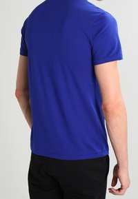 Person sedd bakifrån iförd en enfärgad kungsbla t-shirt med kort ärm och svarta byxor mot en neutral bakgrund.