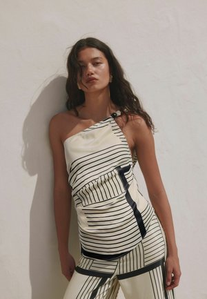 Mladá žena s dlhými tmavými vlasmi má na sebe bezrukávový, jedno rameno odhaľujúci krémový outfit s čiernymi geometrickými pruhmi, stojaca pred jednoduchou svetlou stenou.