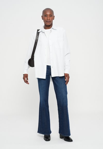 Persona de pie vistiendo una camisa blanca de botones, blusa blanca, jeans acampanados azules, botas negras y un bolso de hombro negro frente a un fondo liso.