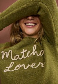 Oliven grøn striktrøje med tekstureret overflade og creamefarvet broderet tekst, der står "Matcha Lovers." Afslappet pasform med rund halsudskæring.