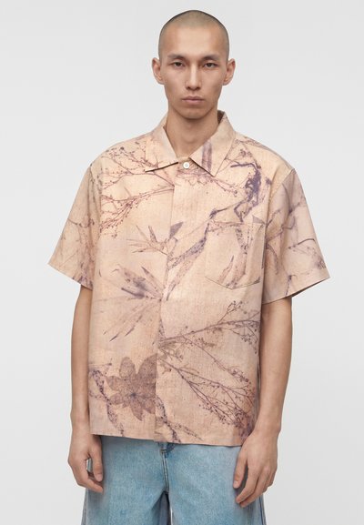 Άνδρας που φοράει μπεζ πουκάμισο με κοντά μανίκια και ξεθωριαστό βοτανικό σχέδιο και ανοιχτό μπλε τζιν, στέκεται μπροστά σε απλό φόντο.