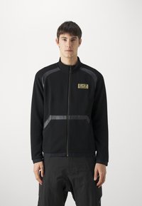 Giacca nera con zip, realizzata in tessuto texturizzato, con accenti grigi e logo EA7 sul petto. Include tasche laterali e un colletto alto.