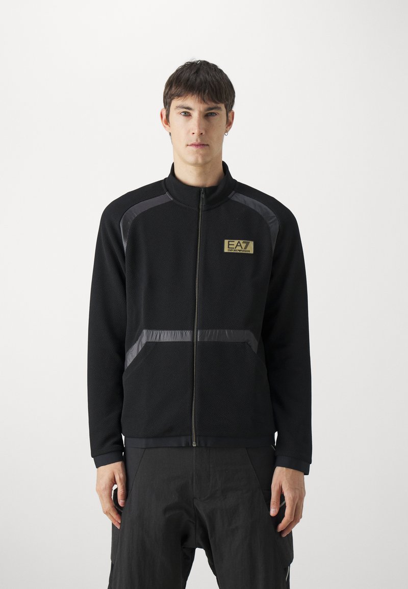 Giacca nera con zip, realizzata in tessuto texturizzato, con accenti grigi e logo EA7 sul petto. Include tasche laterali e un colletto alto.