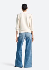 Hanorac de culoarea crem cu mâneci bufante trei sferturi, asortat cu blugi denim bleu deschis cu picioare largi și buzunare în spate.