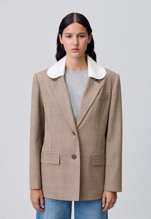 Blazer à carreaux marron avec un col blanc, deux poches avant et une fermeture à un bouton. Porté sur une chemise grise, assorti avec un jean bleu.