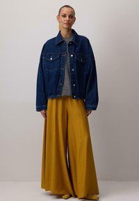 Džínová bunda s knoflíkovým zapínáním, oversized střih, dvě náprsní kapsy. Šedý top pod ní, v kombinaci s širokými žlutými kalhotami a odpovídajícími botami.