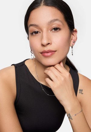 Silberne Ketten-Halskette und Ohrringe. Das Modell trägt ein schwarzes ärmelloses Oberteil. Der Schmuck hat zarte Glieder und hängende Akzente. Der Hautton ist hell.