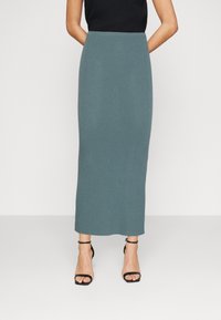 Jupe longue à côtes de couleur bleu-vert atténuée, fabriquée dans un matériau doux. Elle présente une taille ajustée et un design longueur cheville, associée à des sandales noires à lanières.