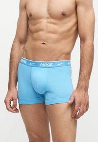 Torse masculin et jambes supérieures portant des boxers Nike bleu clair avec logo sur la ceinture, debout devant un fond clair uni.