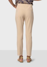 Pantalon beige avec une coupe ajustée, des poches latérales et une taille élastique, assorti de sandales dorées. La texture semble lisse.