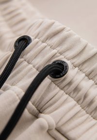 Gros plan d'une ceinture élastique beige avec un cordon noir passant à travers des œillets métalliques noirs sur le tissu.