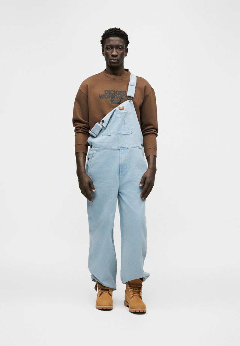 Mann trägt hellblaue Jeans-Latzhose mit einem festgemachten Träger, braunes Sweatshirt und beige Arbeitsstiefel, steht vor einfachem weißem Hintergrund.