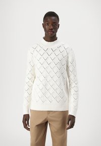 Maglione bianco lavorato a maglia con un motivo a diamante, colletto e polsini a coste. Abbinato a pantaloni beige.
