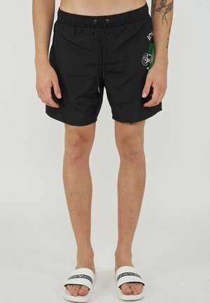 Modèle masculin portant un short de bain noir à cordon avec un imprimé latéral, des sandales Emporio Armani blanches et des tatouages visibles sur les bras.