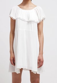 Vestido blanco texturizado con cuello redondo, volantes en capas y ribete de pompones en las mangas. Silueta en A con la parte trasera ligeramente más larga.