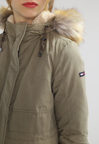 Žena v khaki parce s kapucí s kožíškem a výšivkou loga Tommy Hilfiger na horním rukávu.
