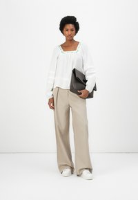 Witte blouse met groene geborduurde halslijn, gecombineerd met beige wijde broek en witte sneakers. Houdt een donkerbruine clutch bij zich.